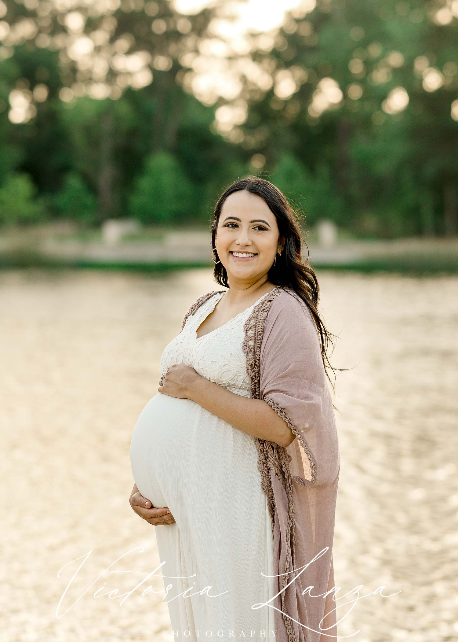 pregnant woman wearing white lace dress posing for a maternity photo session standing behind a lake at sunset