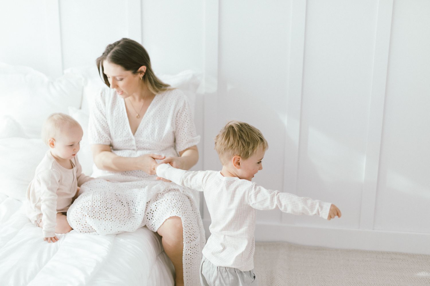 Mother and children share joyful moments together on white bedding in bright minimalist setting.