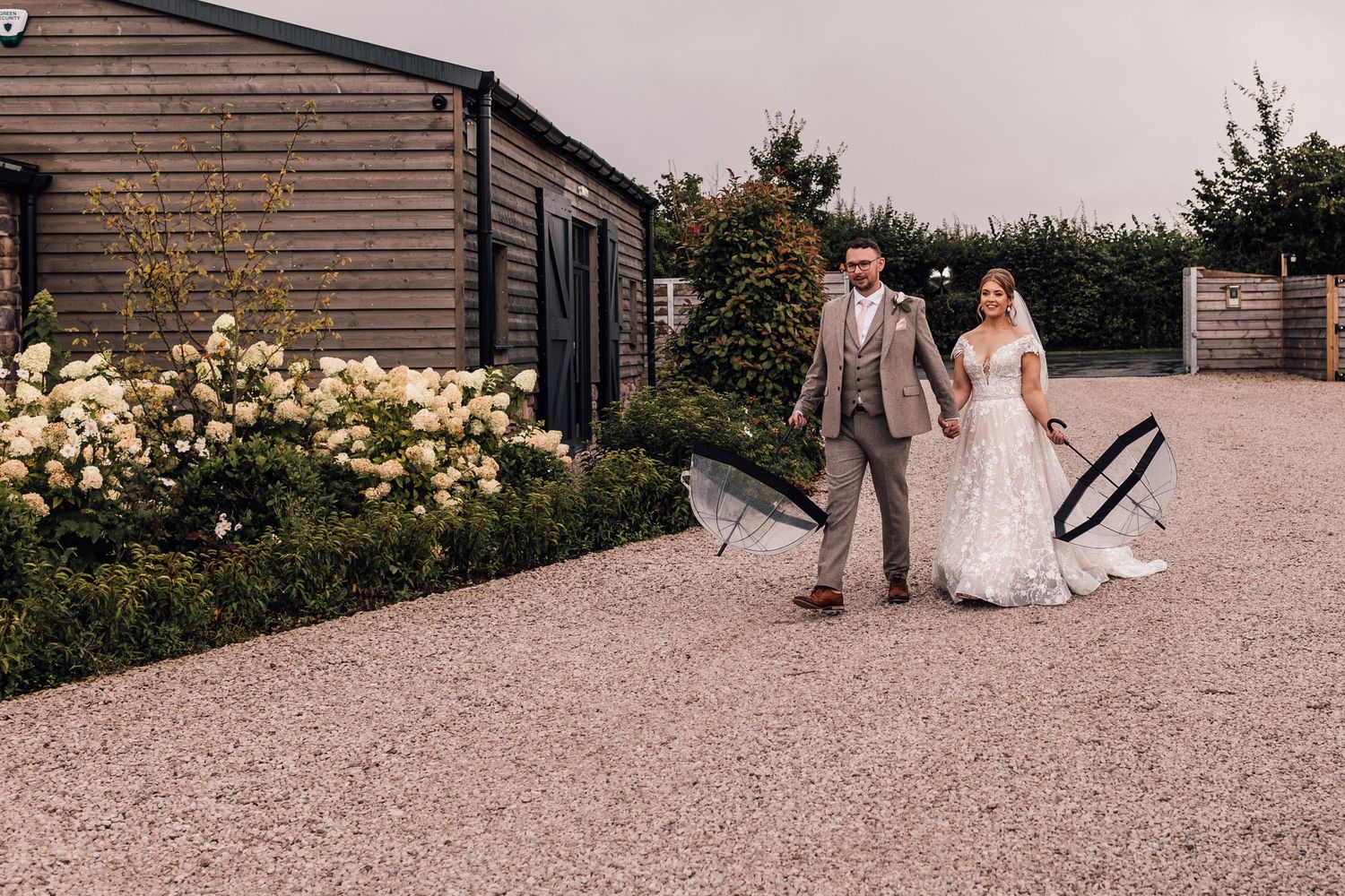 Couple walks along gravel path beside wooden building and blooming garden on wedding day.