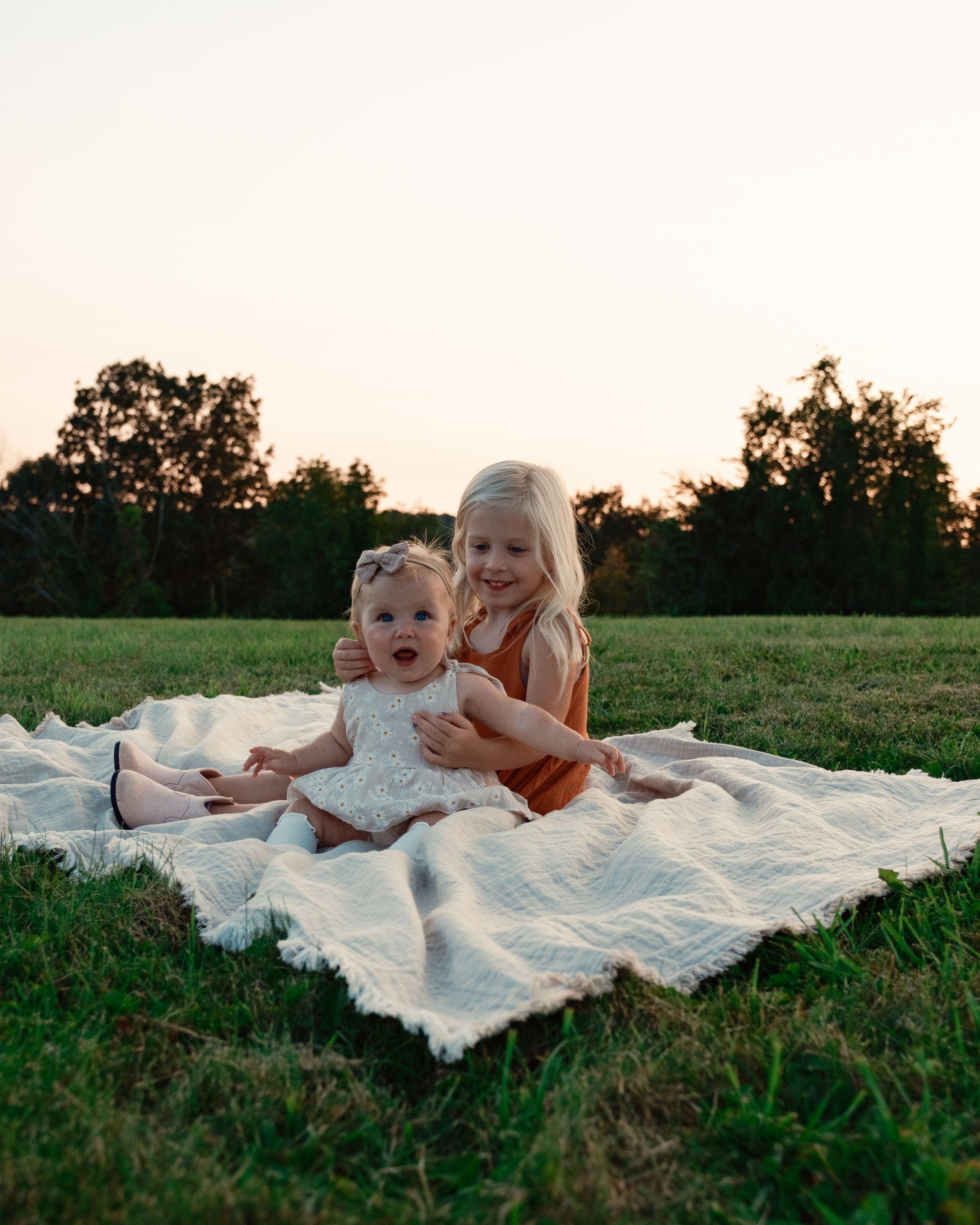 a blonde girl sitting with a baby on a white blanket outdoors