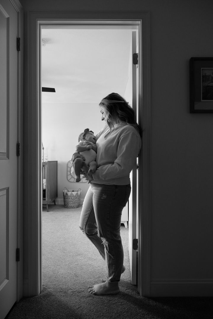 mom wearing jeans standing in doorway of nursery holding newborn