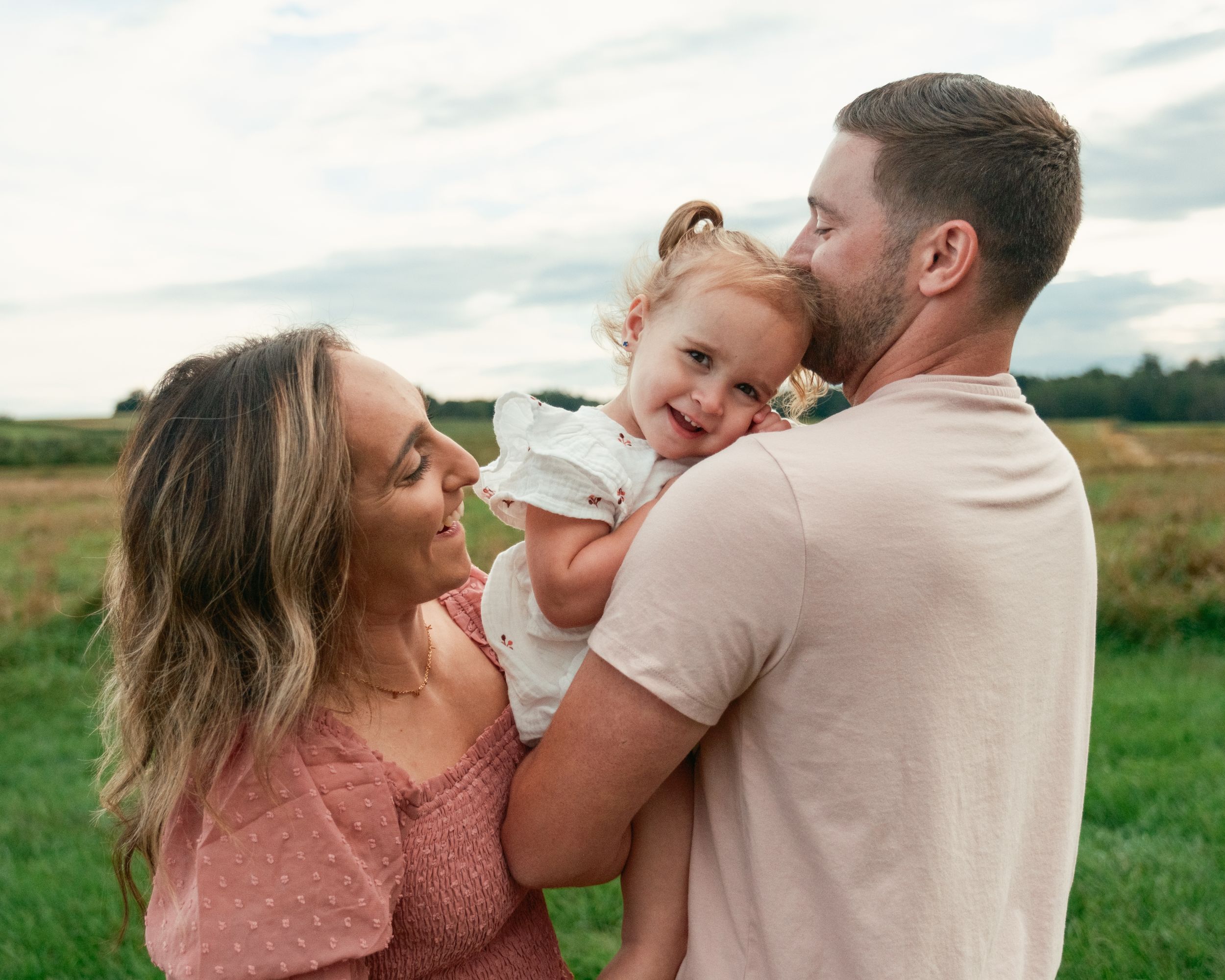 Dad holding toddler girl with mom laughing standing close together in a field