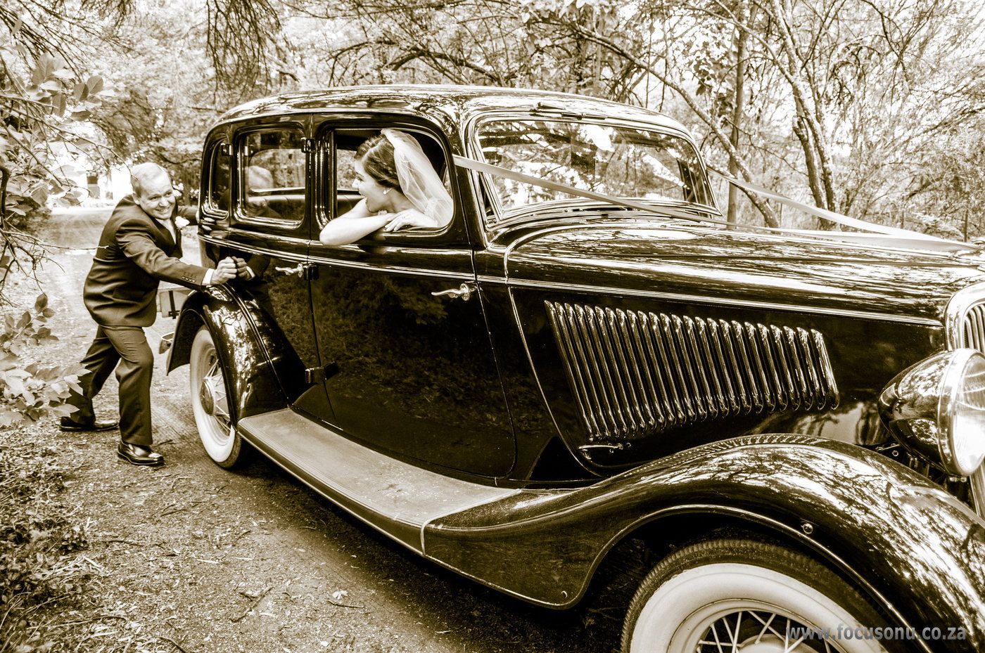 Groom pushing an old classic car while the bride encourage him