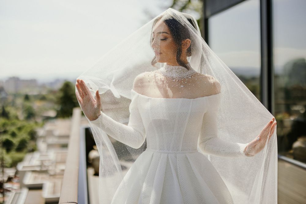 Bride in long-sleeved white gown and veil standing on balcony with city view.