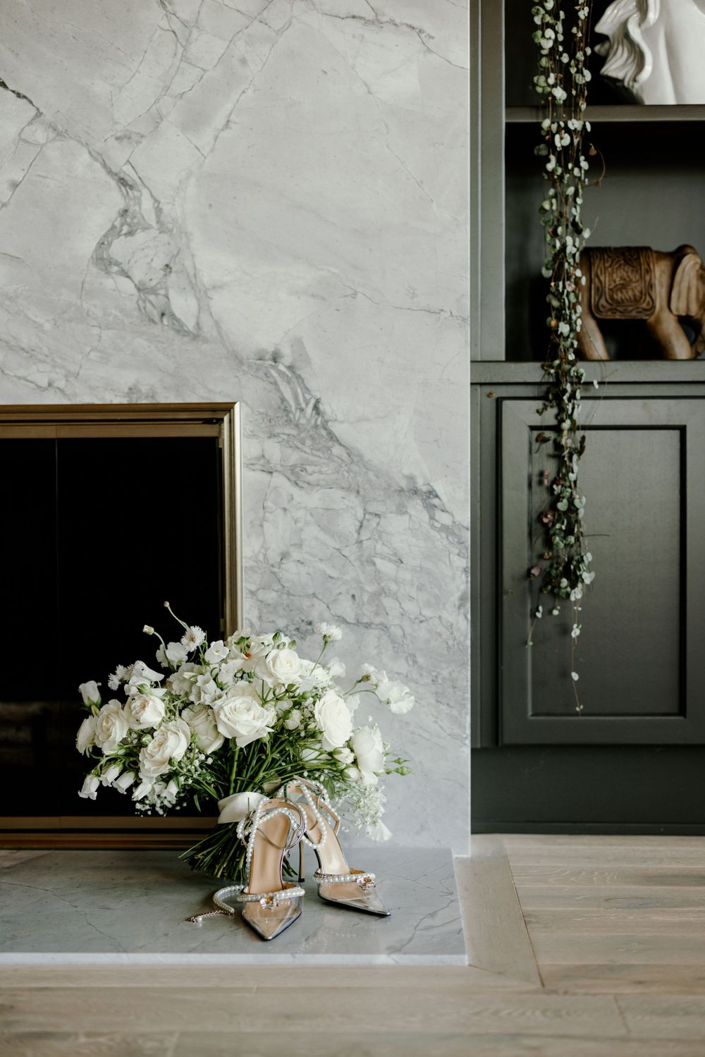 Wedding details featuring elegant white floral arrangements and bridal shoes against marble fireplace.