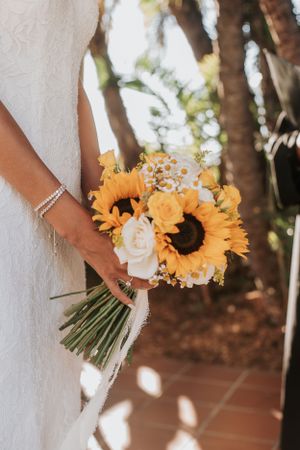 Bridal bouquet with sunflowers and white roses held against white wedding dress.