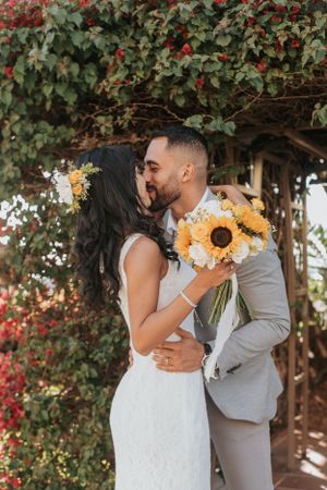 Newlyweds share a romantic kiss under a flowering archway with vibrant sunflower bouquet.