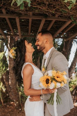 Romantic wedding portrait under wooden pergola with sunflower bouquet and natural lighting.