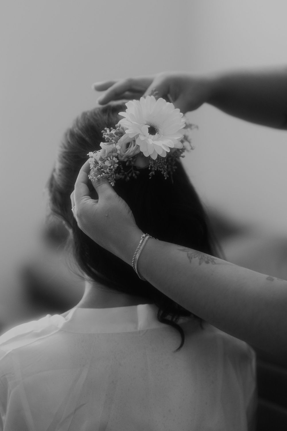 Black and white photo of hands placing a white flower in someone's hair during wedding preparations.
