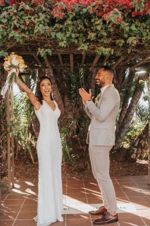 Wedding couple celebrating under pergola with red flowers, bride holding bouquet triumphantly.