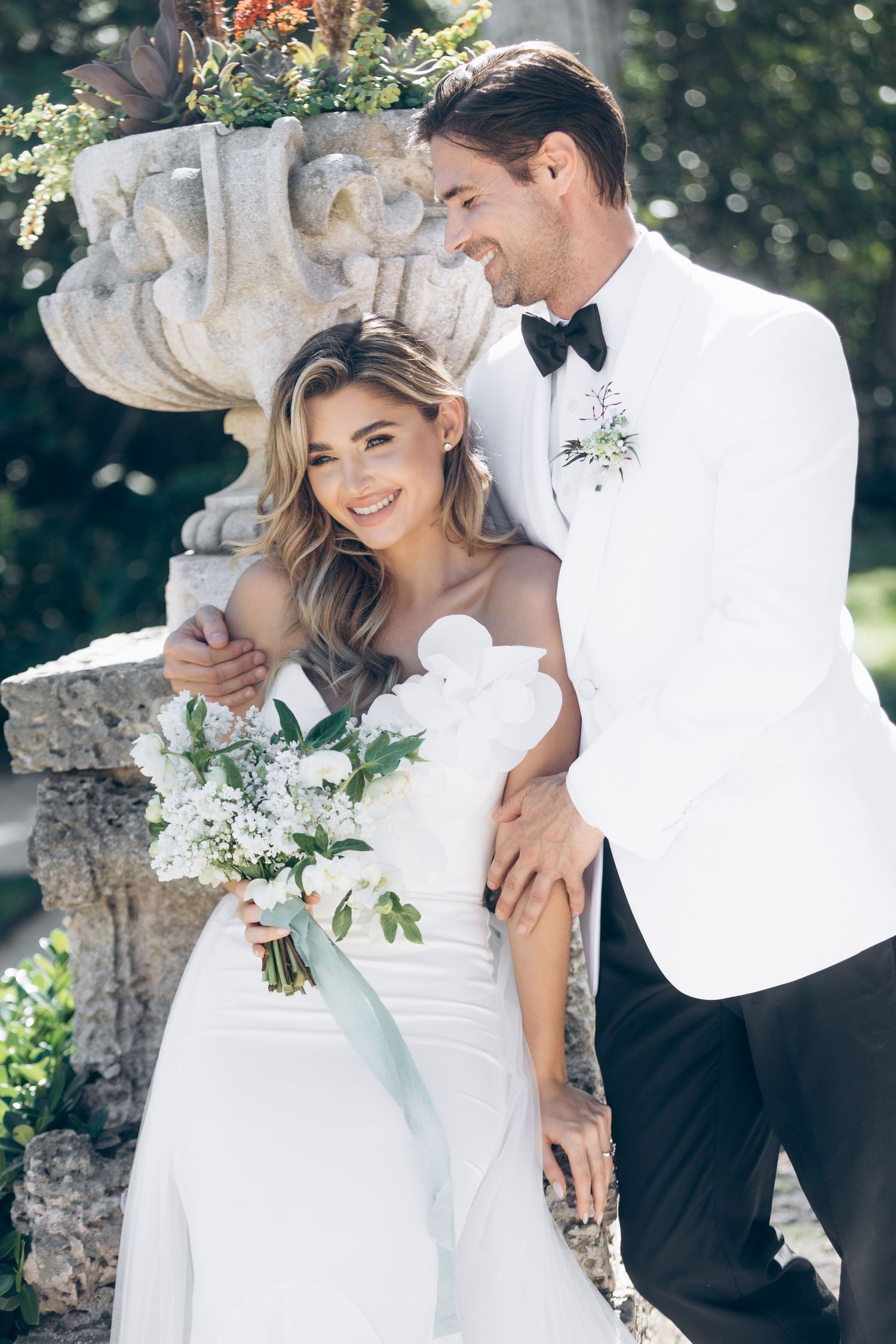 An elegant wedding portrait captured in a garden setting with ornate stone architecture.