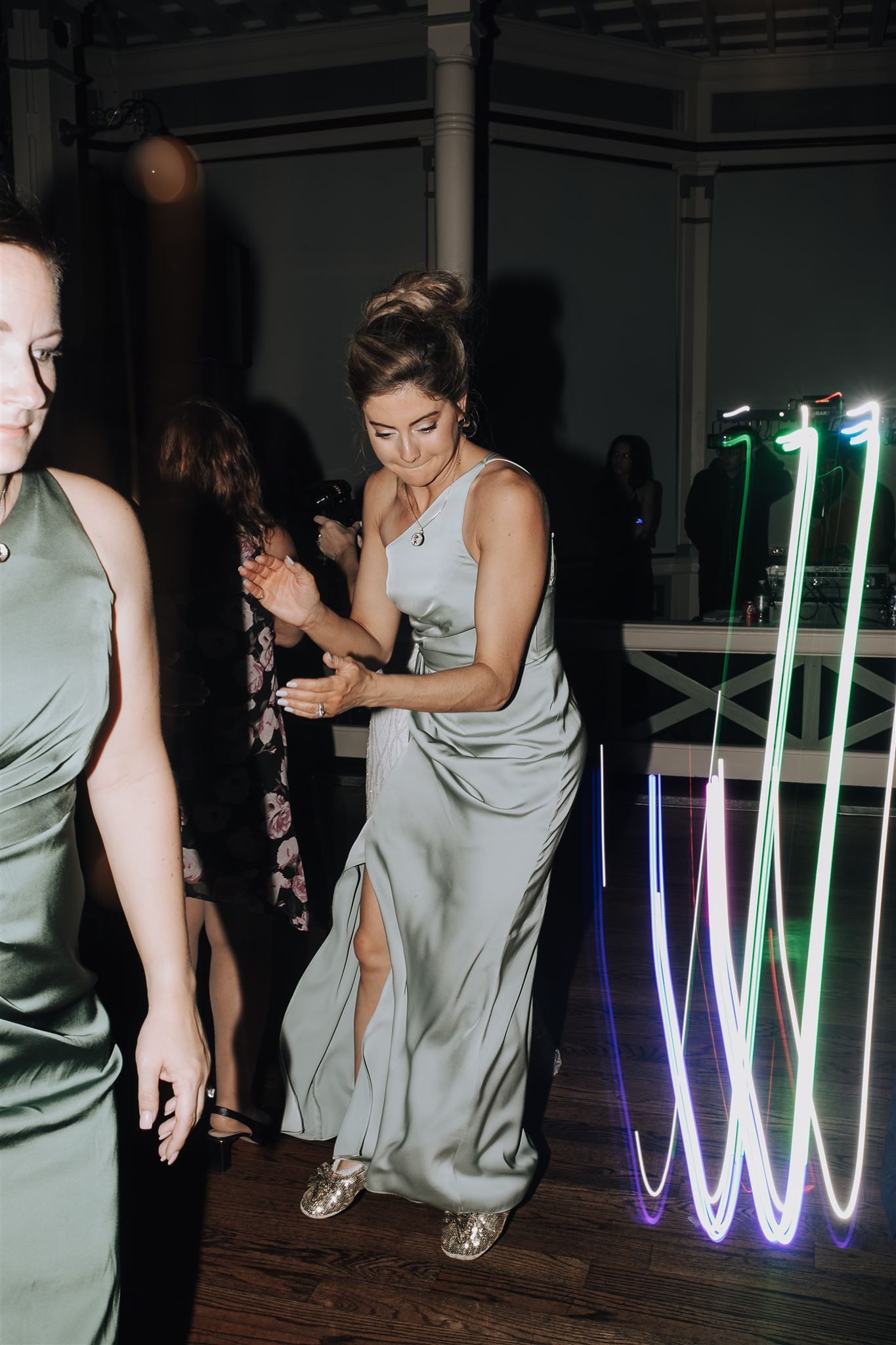 A person in a light gray dress dances near illuminated neon lights at a party.