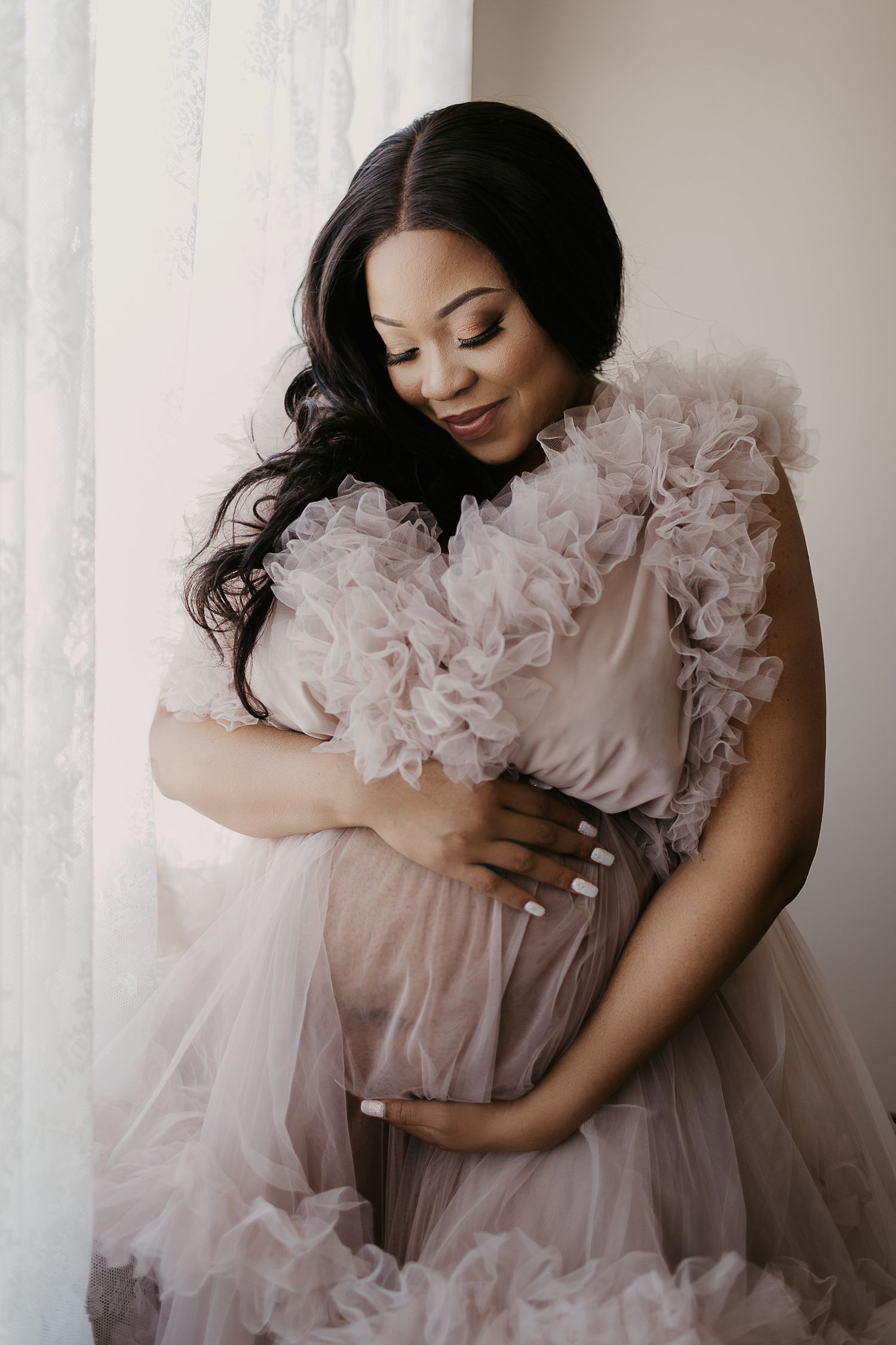 Maternity portrait of woman in flowing tulle dress posing by window.