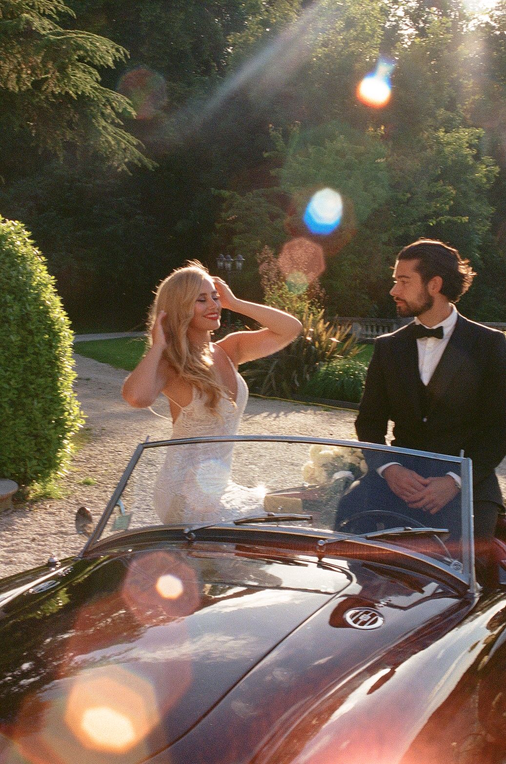 A carefree couple in wedding attire sit in a vintage convertible car surrounded by sunlit gardens.