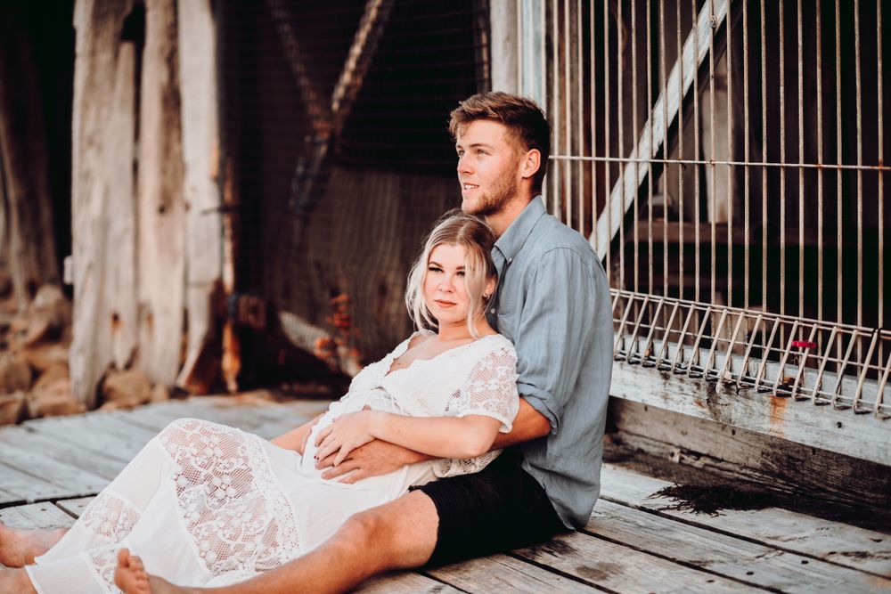 Couple embraces on rustic wooden deck during maternity photoshoot in casual attire.