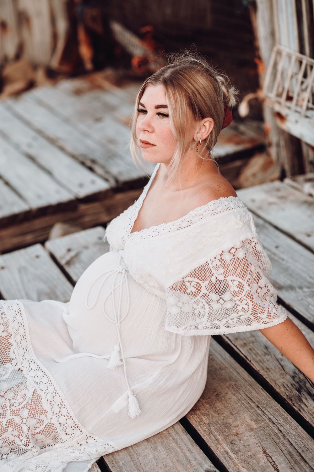 Expectant mother in white lace maternity dress sits on wooden deck during sunset photoshoot.