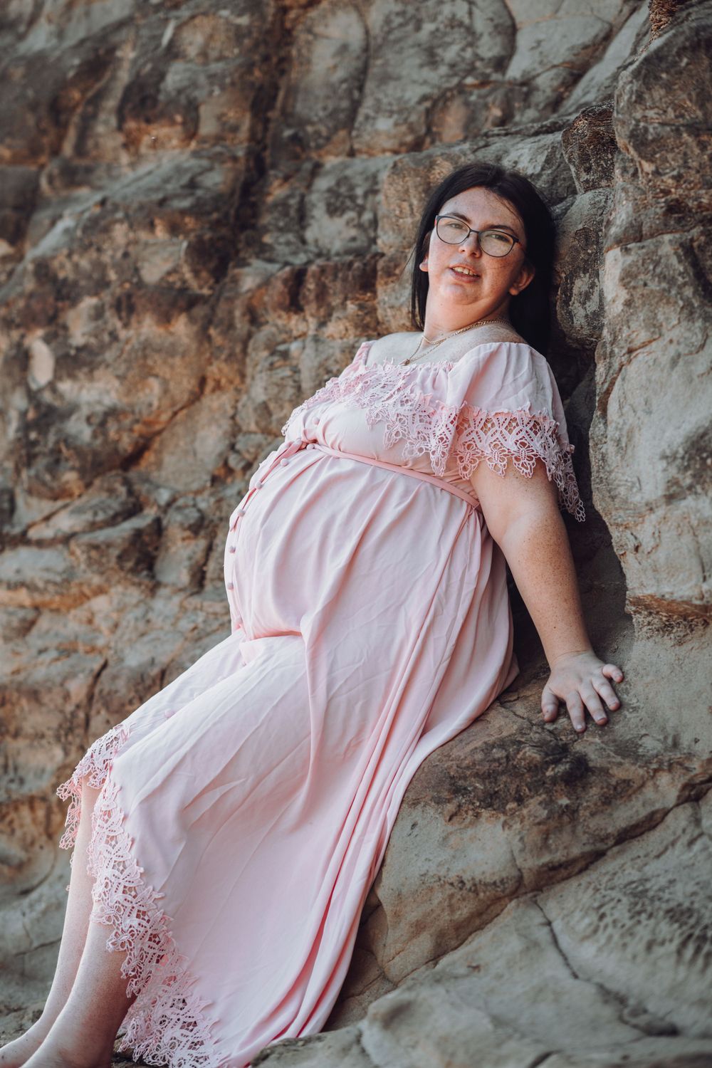 Pregnant woman in pink lace dress poses against textured rock wall during maternity shoot.