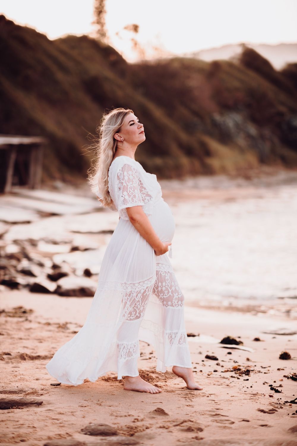Pregnant woman in flowing white lace dress poses on beach at sunset.