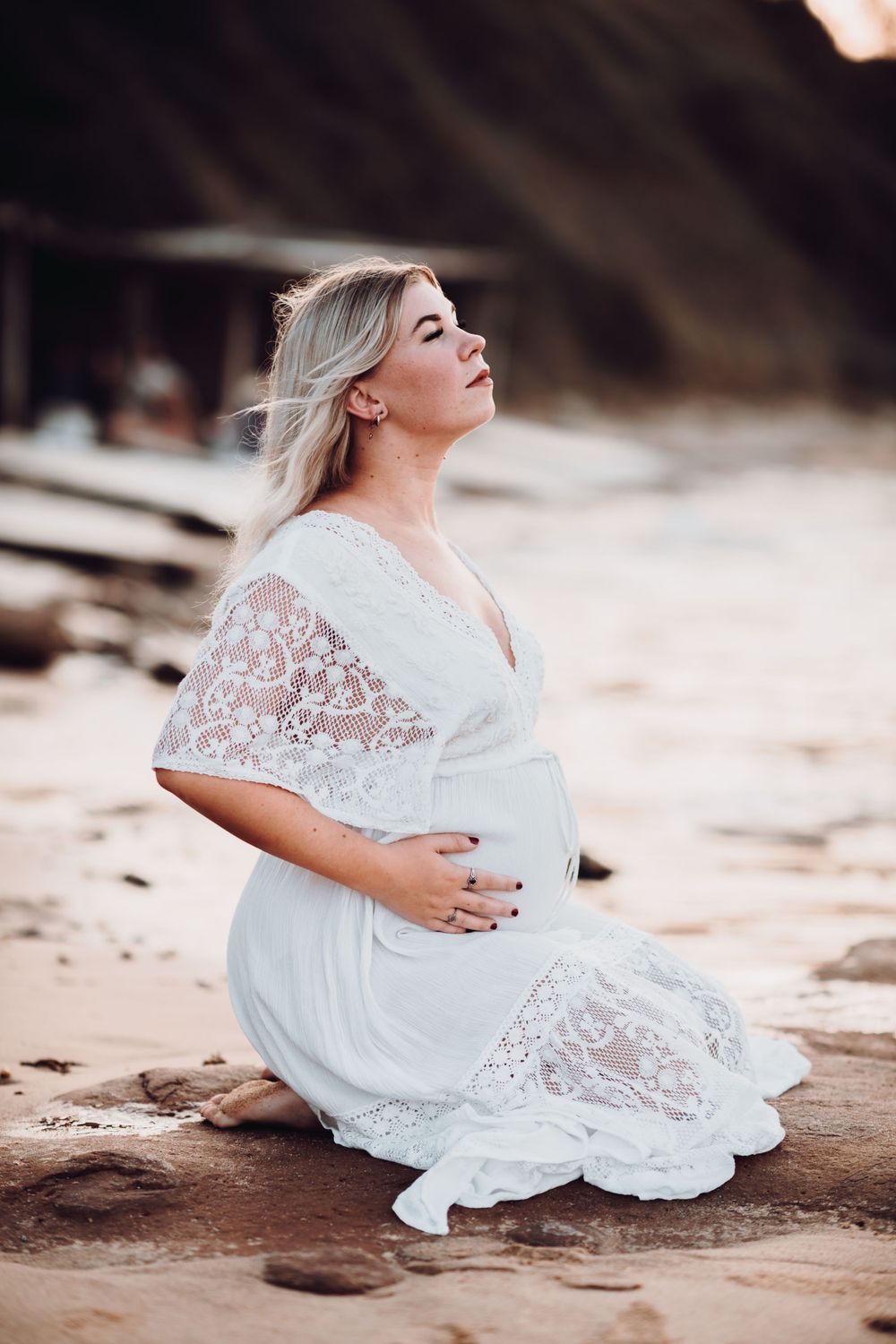 Maternity beach portrait of woman in white lace dress sitting on sandy shore.