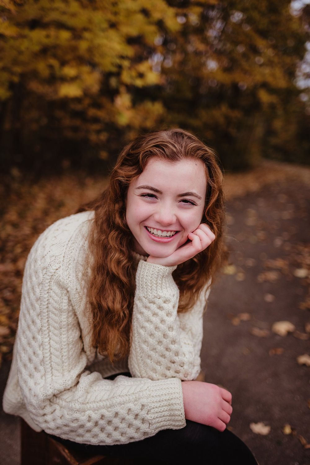 Person in cream sweater poses cheerfully on autumn path with golden trees in background.