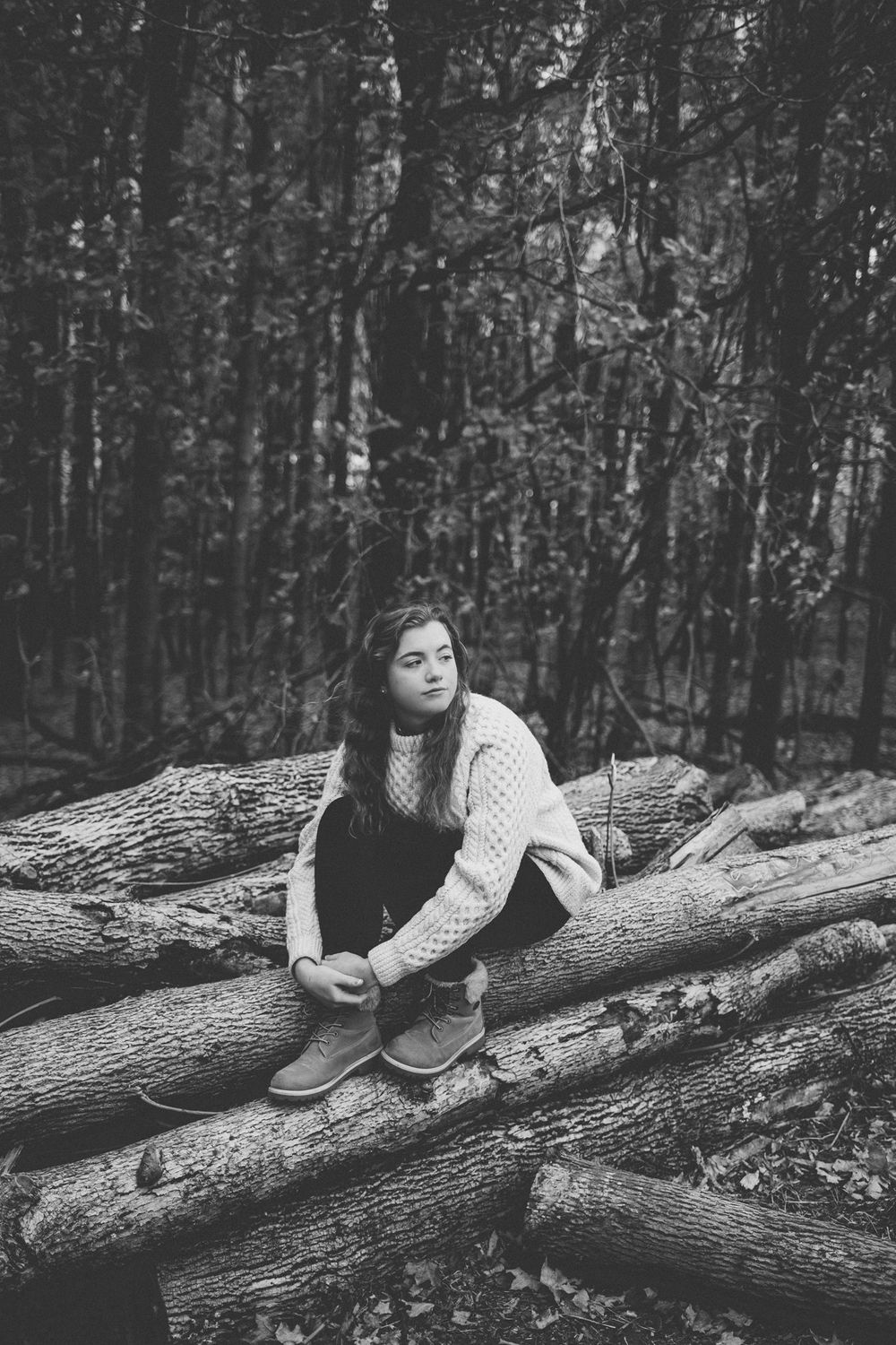 A moody black and white portrait of a person sitting on a pile of logs in a forest setting.