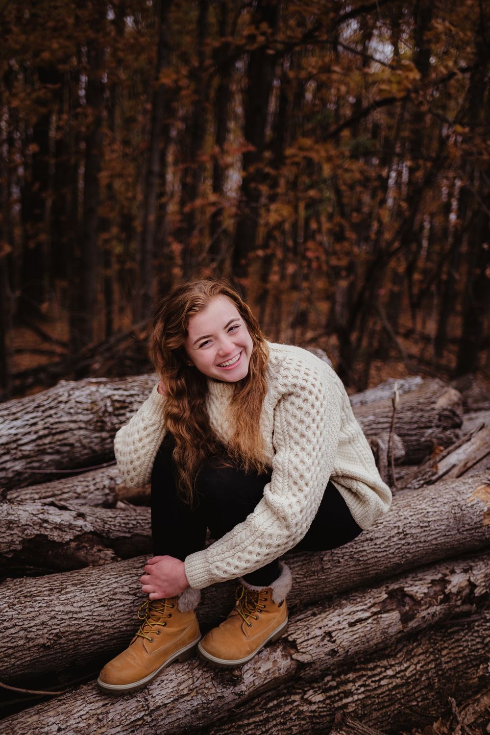Person in cream sweater sitting on fallen logs in autumn forest.