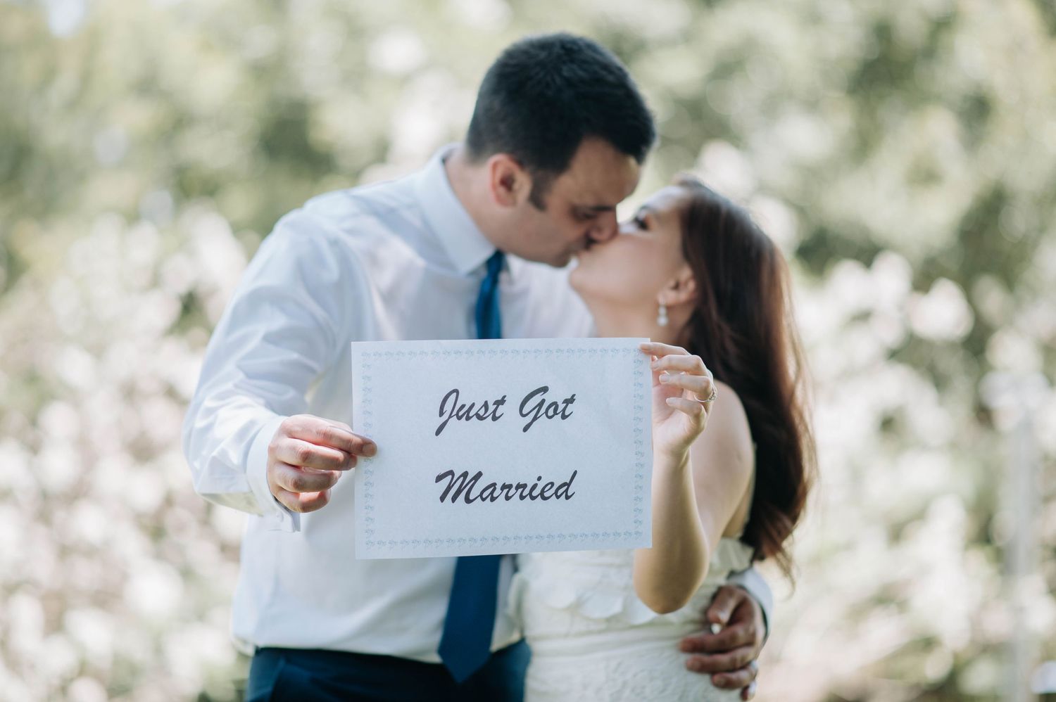 Wedding couple sharing a kiss while holding a 'Just Got Married' sign outdoors.