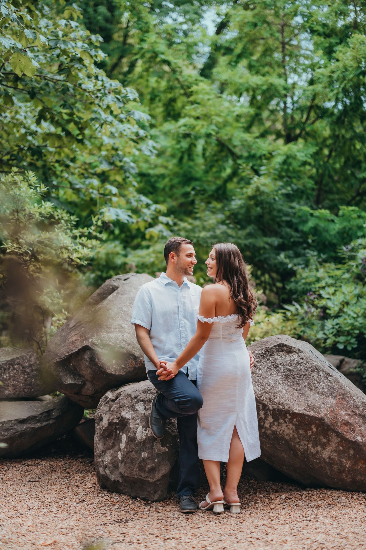 Couple holding hands and gazing at each other in white outfits among large rocks in a lush green park.