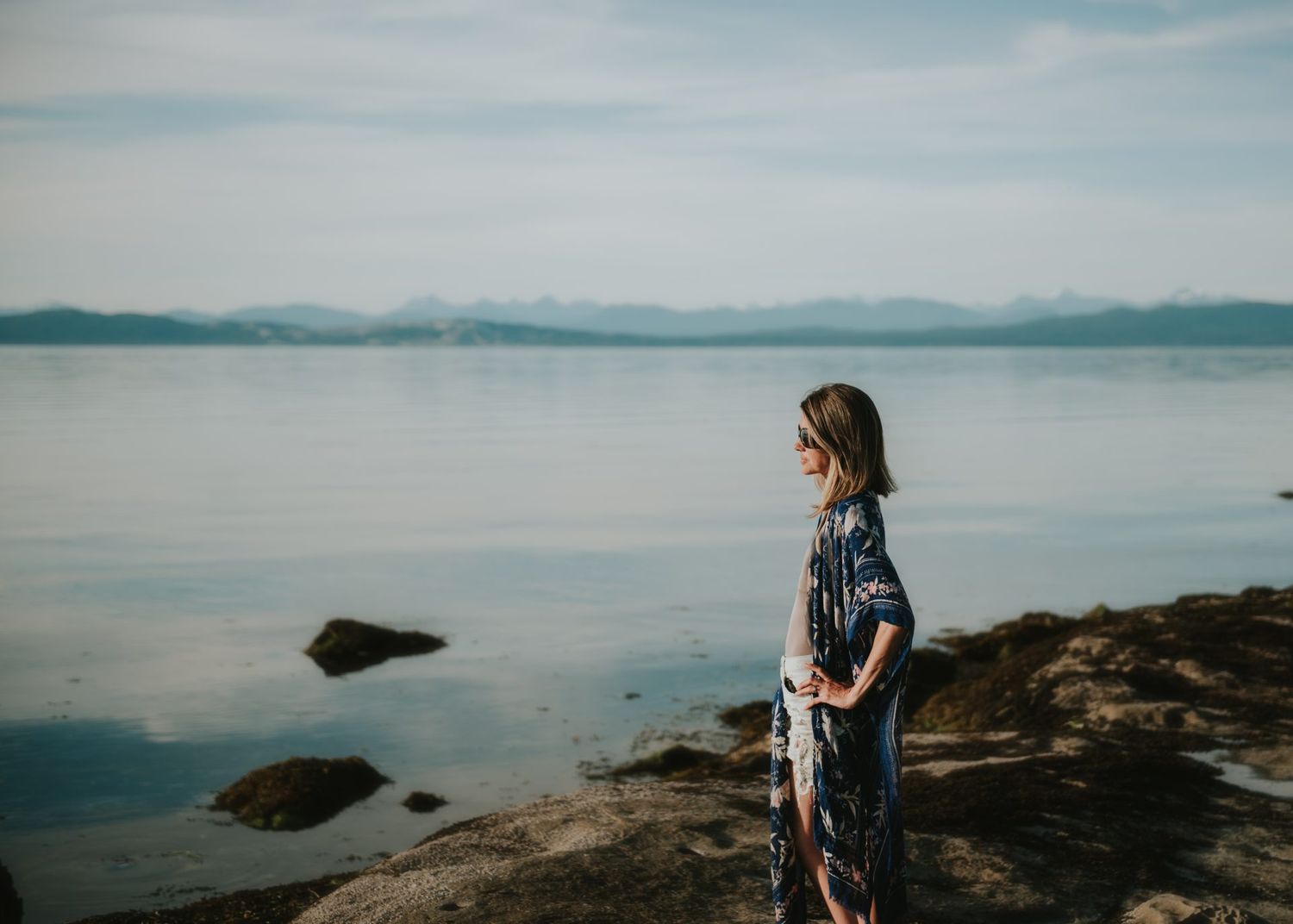 Person stands contemplatively on rocky shore overlooking calm mountain lake.