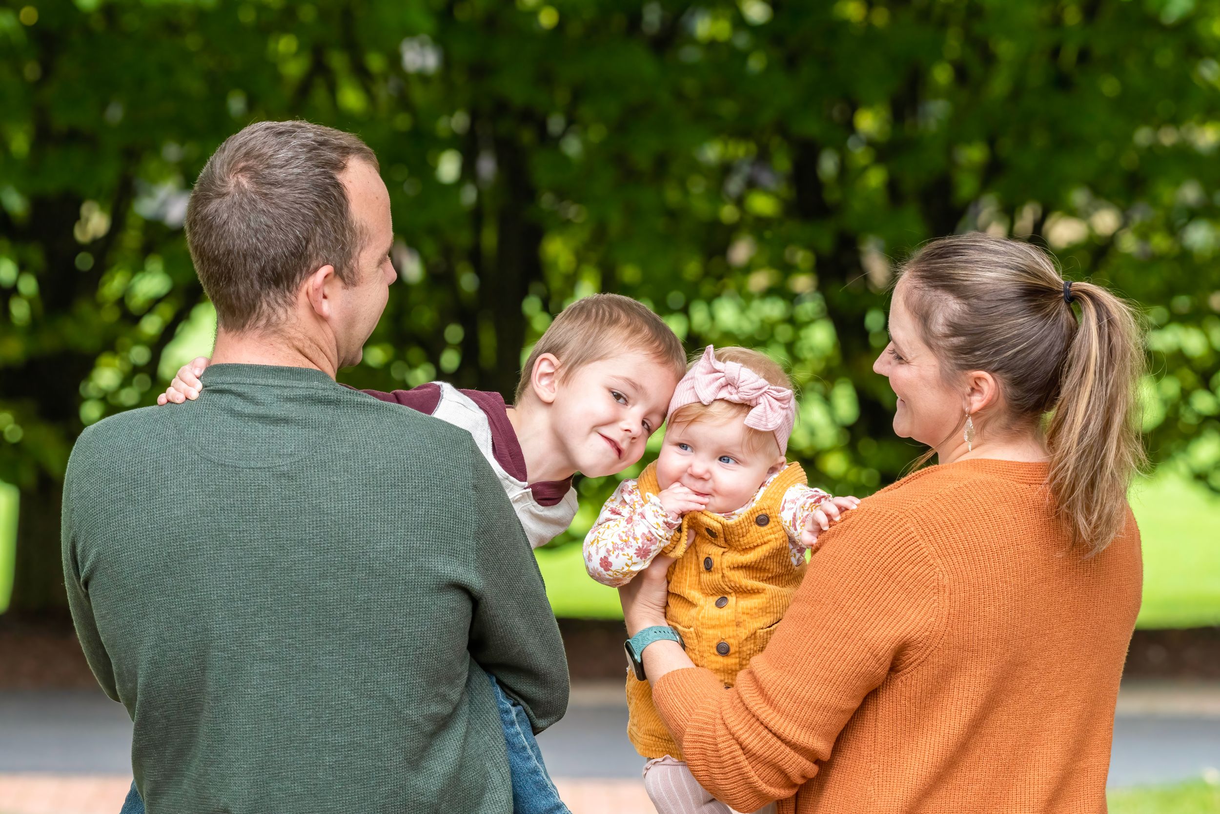 A family in green and orange sweaters shares a playful outdoor moment together in a park setting.