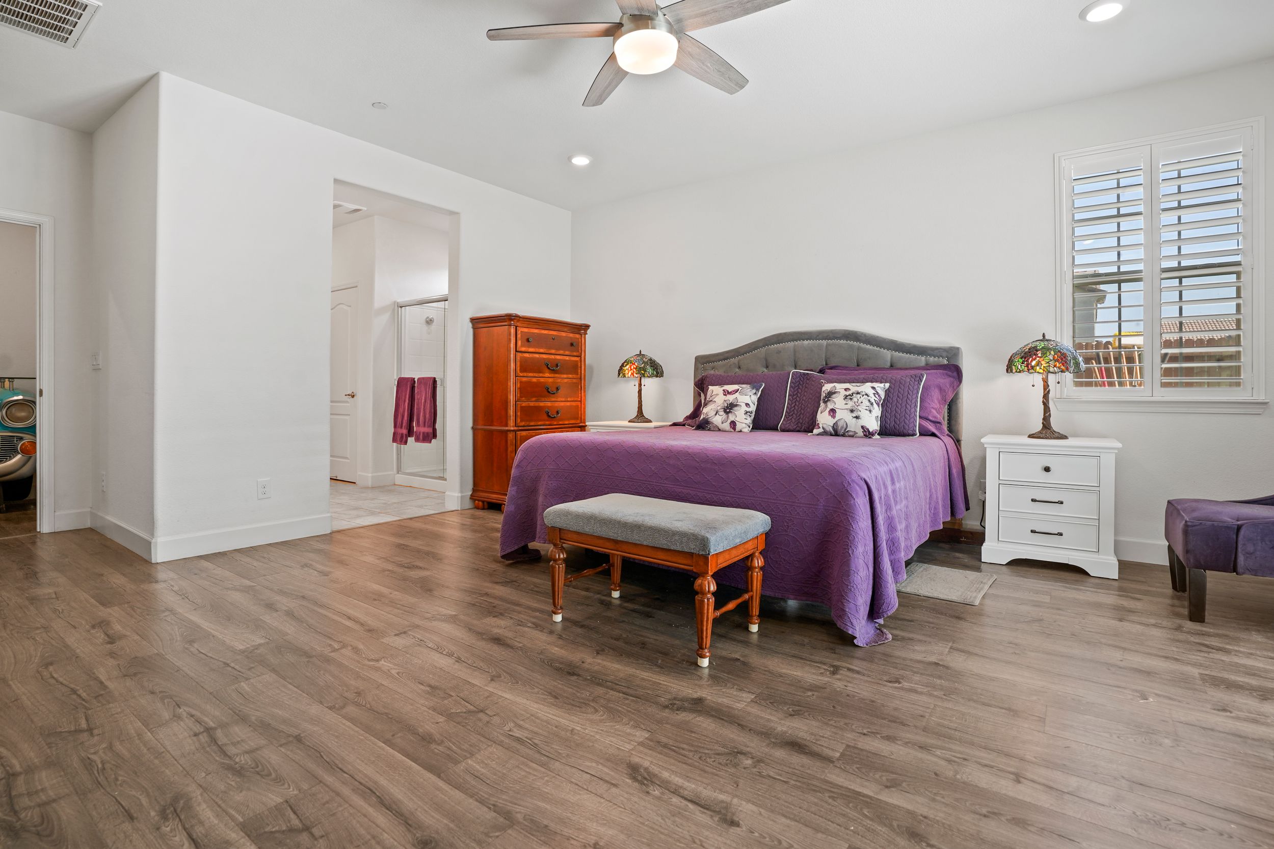 Spacious master bedroom with purple bedding, hardwood floors, and white walls.