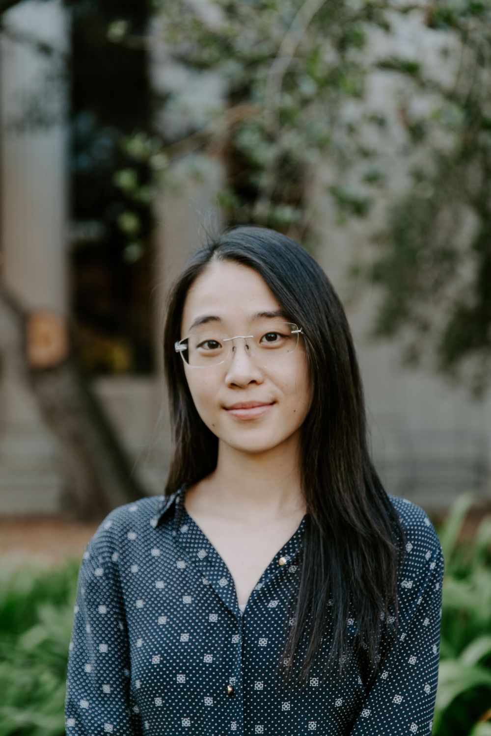 Portrait of a young woman in glasses and polka dot shirt against a natural outdoor background.