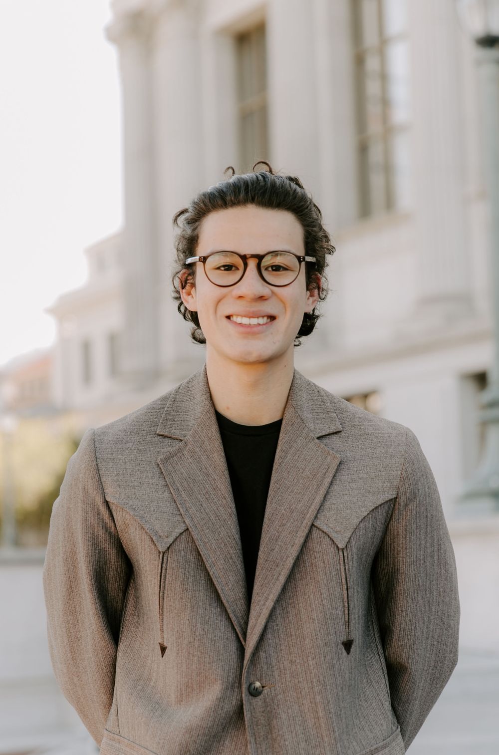 Person wearing a grey blazer and black turtleneck posing outdoors near white columns.