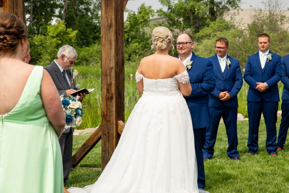 Outdoor wedding ceremony under wooden arch with wedding party in blue suits and mint green dress.