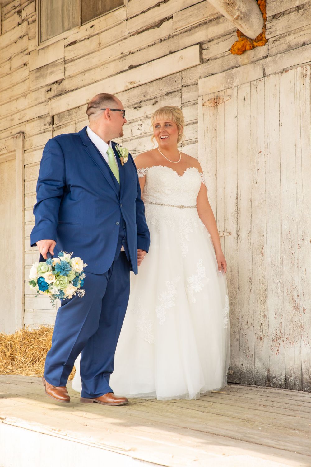 Couple in wedding attire pose together in front of weathered white barn with blue bouquet.