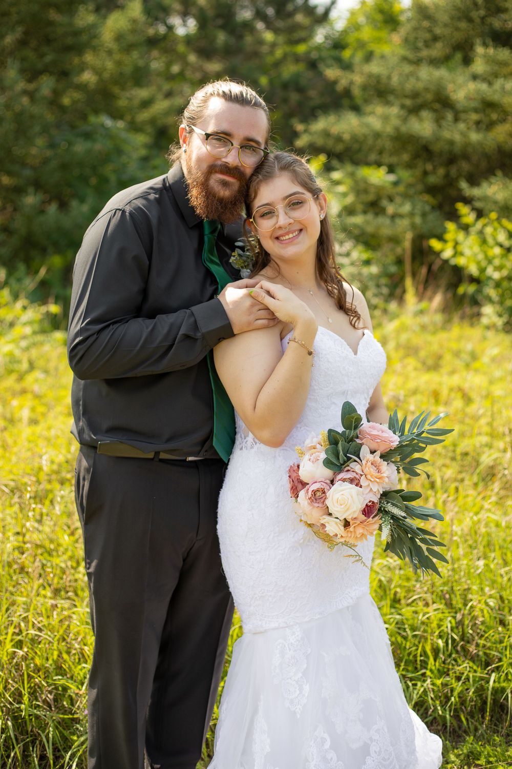 A couple embraces in wedding attire while standing in a sunny meadow.