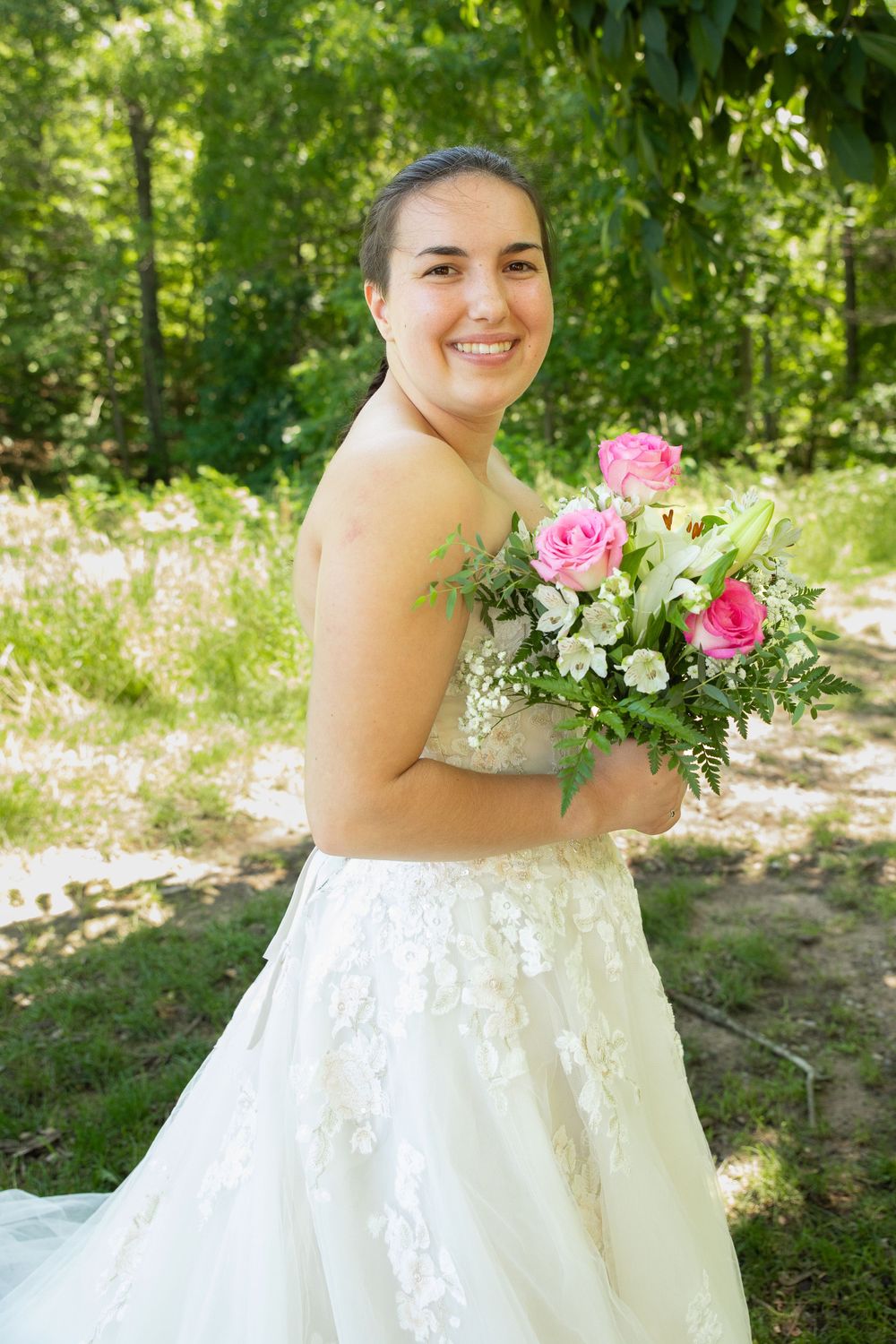 Bride in white lace dress holds pink and white bouquet outdoors.