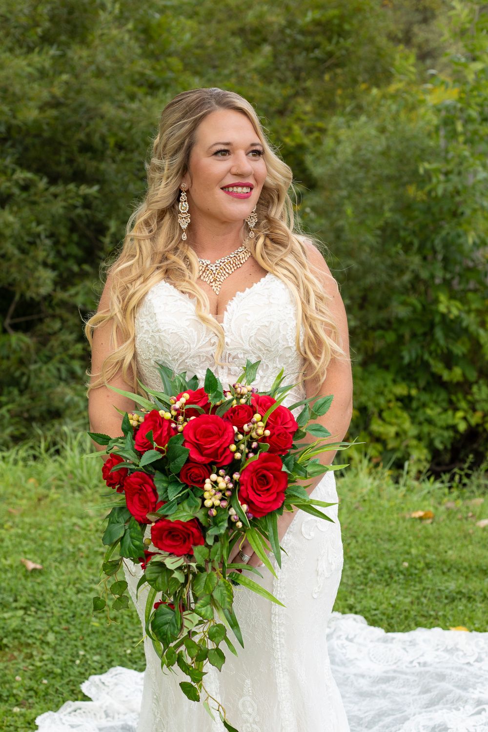 A bride in a lace white dress holds a cascading red rose bouquet against a green garden backdrop.