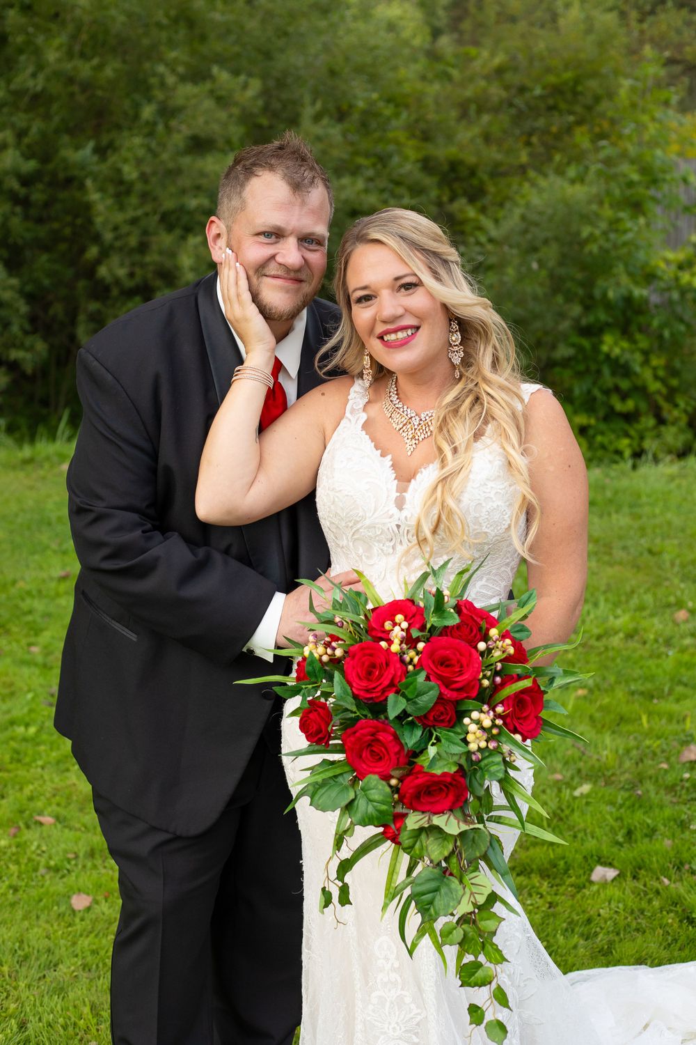 A happy wedding couple poses together outdoors with a vibrant red rose bouquet.