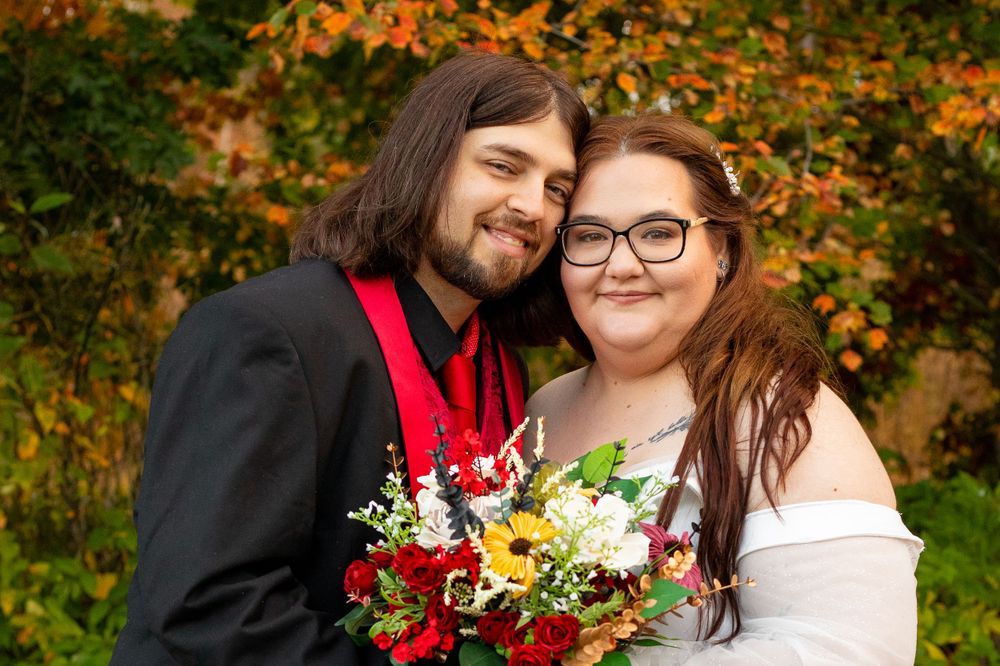A couple in wedding attire smiles together against colorful fall leaves.