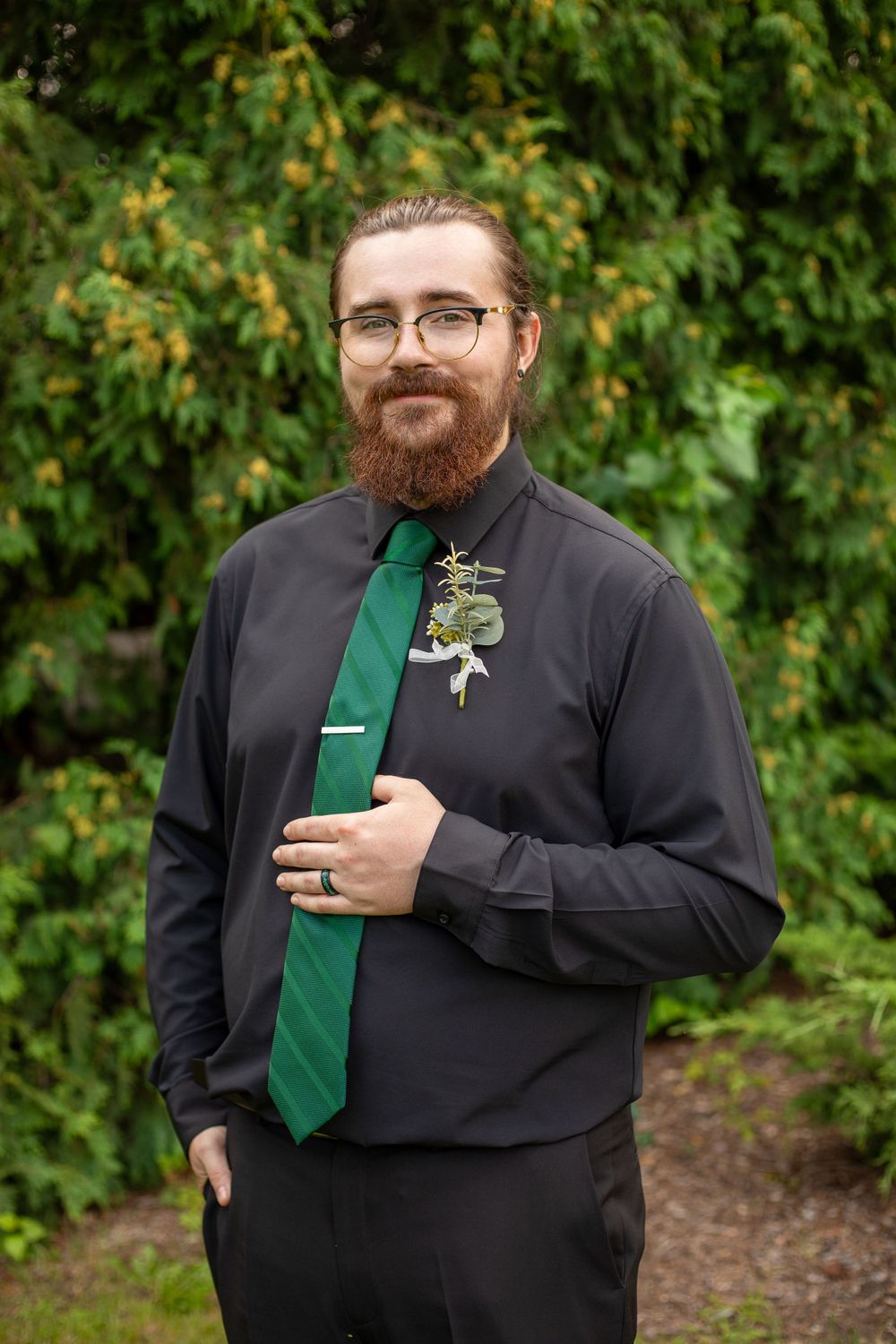 A formal portrait of someone wearing a black shirt, emerald green tie and boutonniere in front of greenery.