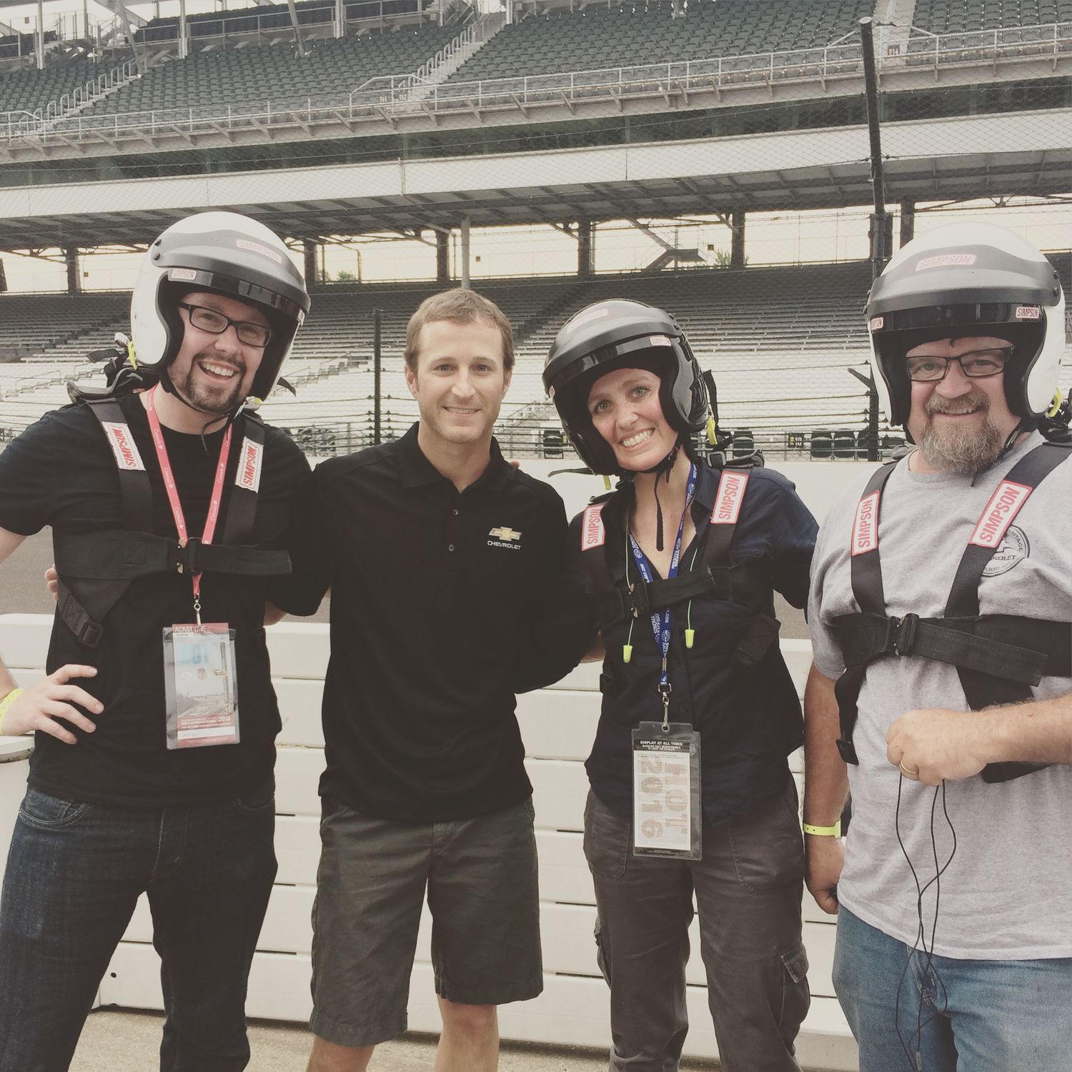 A group wearing racing helmets poses for a photo at what appears to be Indianapolis Motor Speedway.