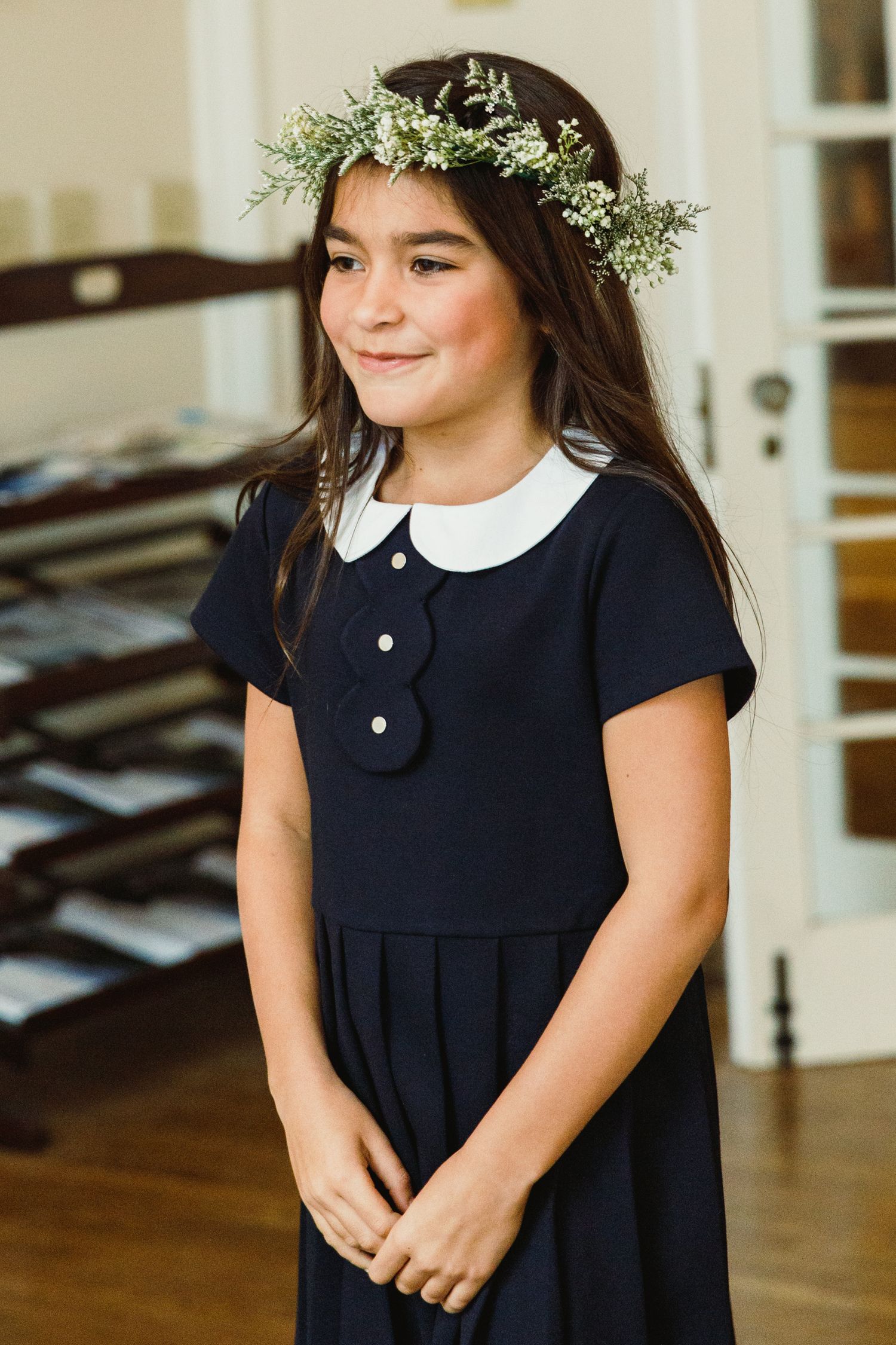 A child wearing a navy dress with white collar and floral crown stands in an elegant indoor setting.