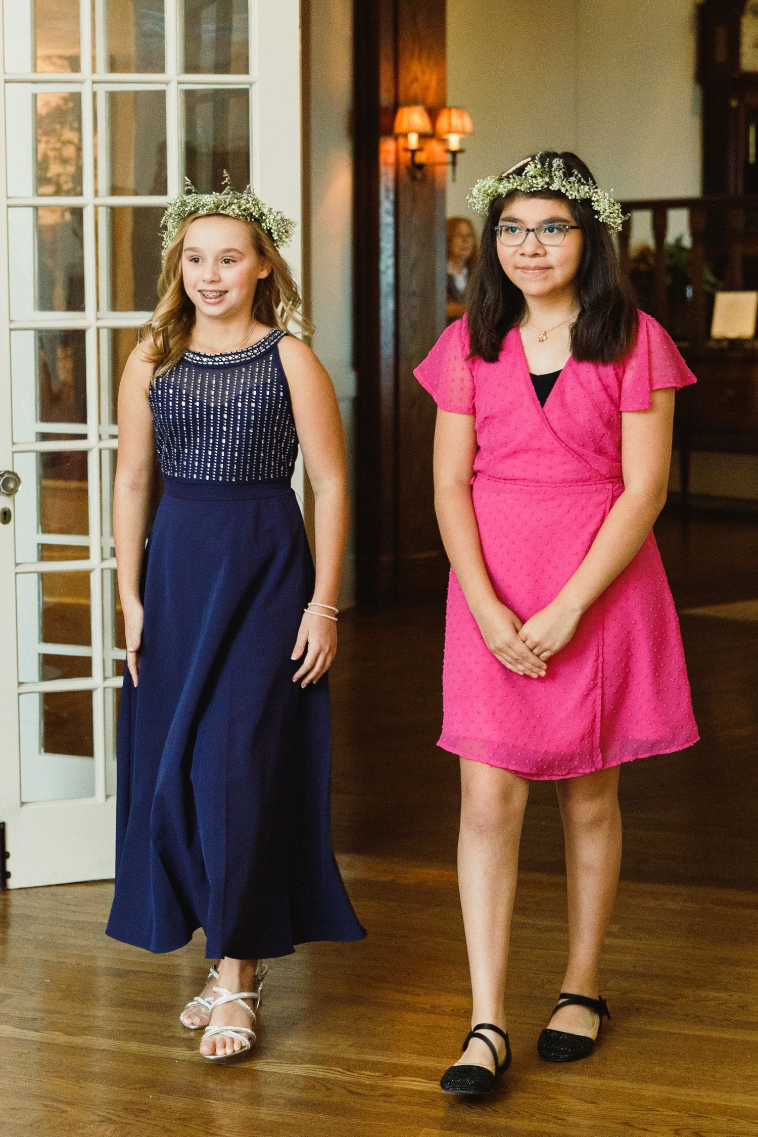 Children in formal dresses stand in a line on hardwood floors at an indoor event.