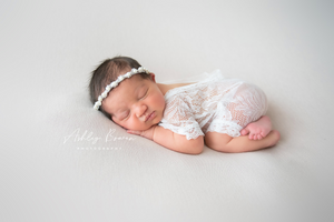 Peaceful newborn baby in white lace romper sleeping on soft white backdrop.