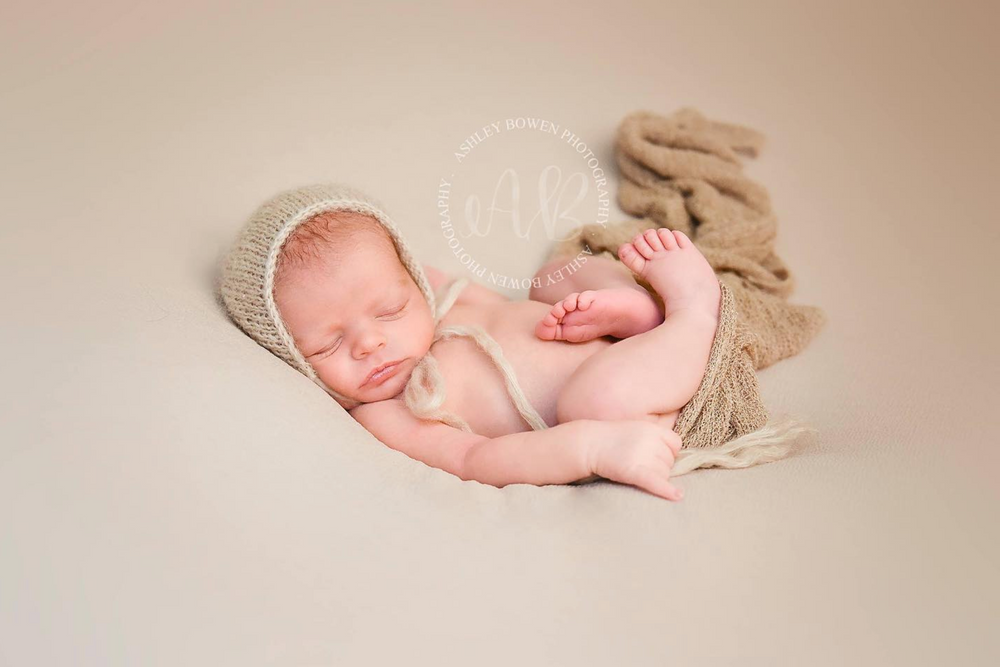 Newborn baby in knit bonnet posed on beige blanket with burlap wrap.