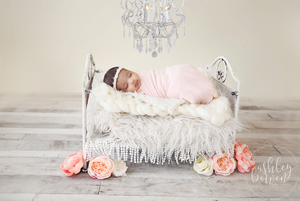 Swaddled newborn in pink wrap on fur-covered miniature bed with pink roses.