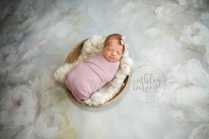 Sleeping newborn wrapped in purple swaddle in wooden bowl on floral painted backdrop.