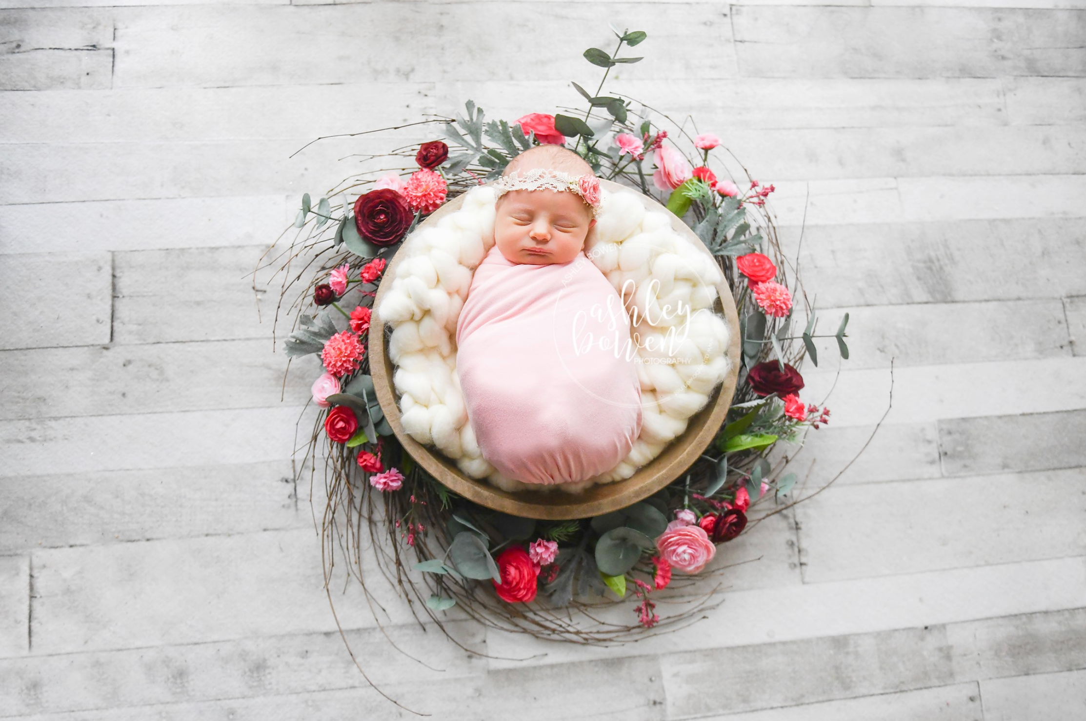 Newborn baby in pink swaddle in wooden bowl surrounded by red and pink flowers.
