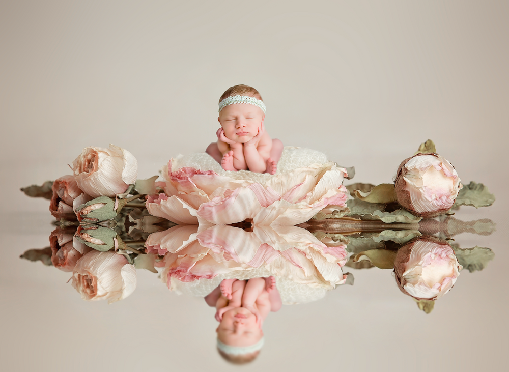 Newborn baby resting chin on hands surrounded by pink roses with mirror reflection on white surface.